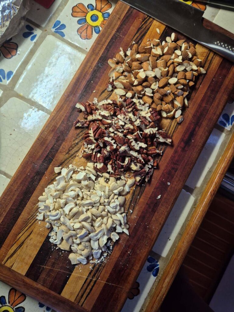 Rectangular dark wood cutting board with 3 piles of roughly chopped raw nuts - cashews, pecans and almonds